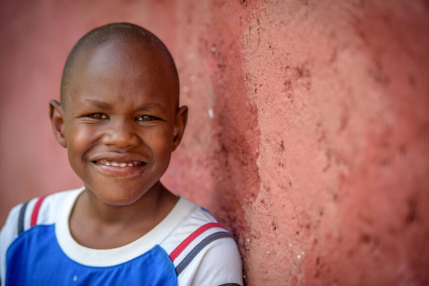 A Kenyan boy leans against a red wall and smiles at the camera.
