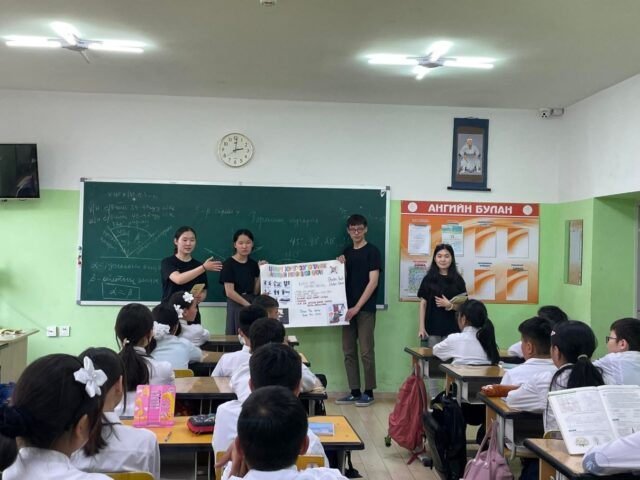 Three students hold a sign at the front of a full classroom.