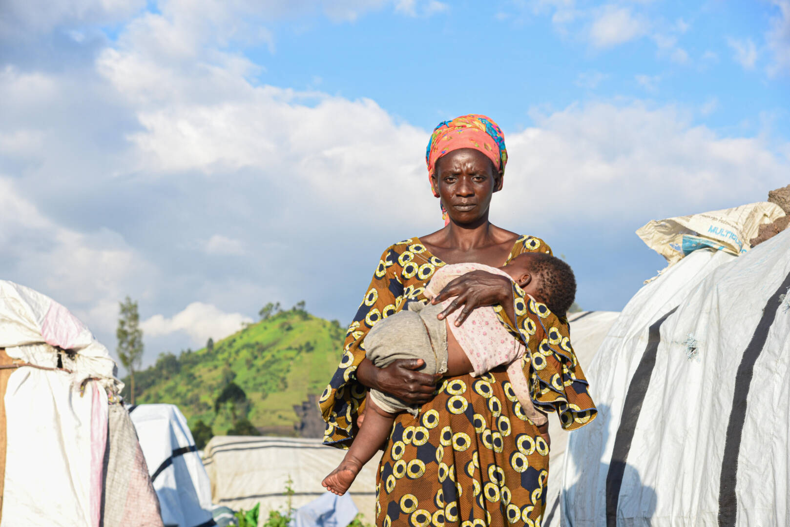 A woman wearing a patterned dress holds a sleeping infant in her arms. She stands between two rows of makeshift tents.