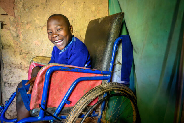 A smiling boy wearing a school uniform sits in a wheelchair.