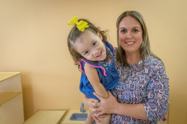 A little girl leans toward the camera smiling while a woman holds her.