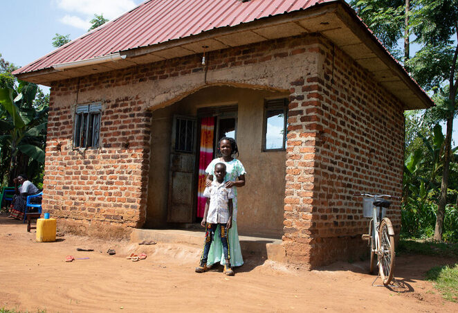 Rome, a World Vision sponsored child, and his mom, Hellen, stand in front of the new house their family was able to build in Uganda as a result of a Special Gift from his sponsor and savings Hellen accrued through her savings group.