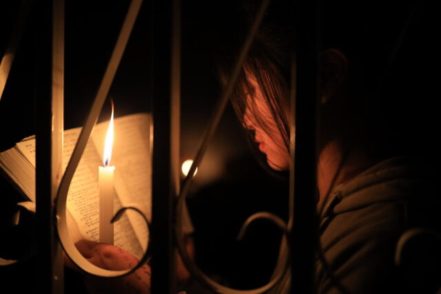 A girl illuminated by candlelight gazes onto the pages of an opened book, viewed from behind ornate iron bars.