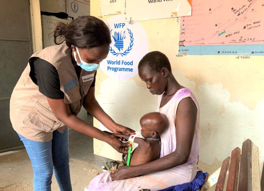At a World Vision-supported facility in South Sudan, a healthcare worker wearing a blue mask, black T-shirt, and brown vest measures the mid-upper arm circumference of a toddler’s left arm for malnutrition. The armband shows red, indicating the boy is severely malnourished. The child sits on his mother’s lap, and she looks at him wearily and intently.