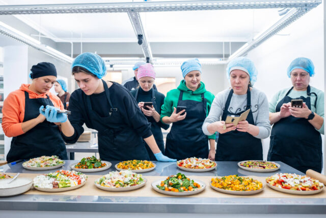 A group of women wearing aprons and hairnets take pictures of dishes of food using their cellphones.
