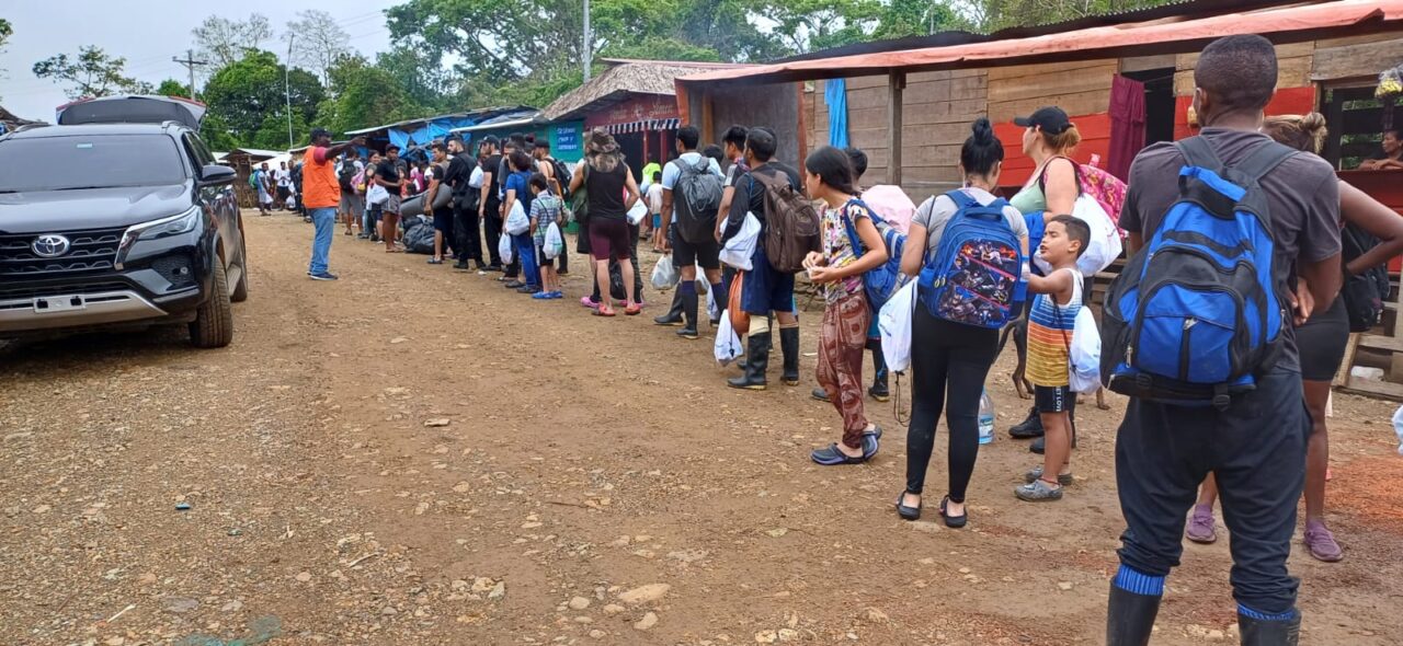 Several dozen people, including children, carry backpacks and form a line in Panama. A man in an orange vest directs them.