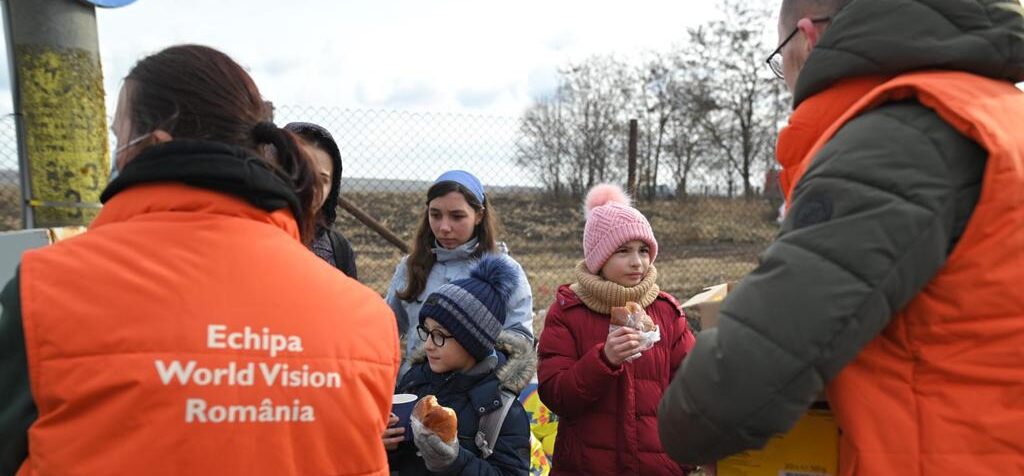 Three children wearing winter coats and hats and holding baked goods face two World Vision staff members in heavy orange vests.