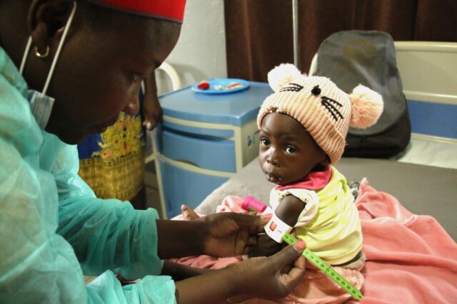 A healthcare worker measures a child's arm for malnutrition; the boy wears a cap with pom-pom ears and a stitched nose and whiskers.