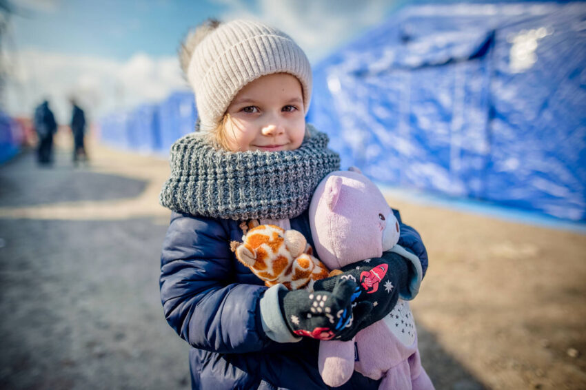 A Ukrainian girl wearing winter clothes and holding two stuffed animals stands outside next to blue tents.