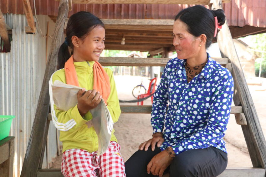 A Cambodian girl in red checkered pants and a bright green jacket sits on outdoor steps as she smiles toward a woman in a blue polka-dotted shirt.
