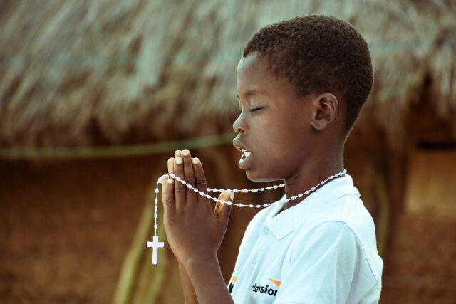 A profile of a boy in a World Vision T-shirt, eyes closed in prayer, with a silver chain and cross in his hands.