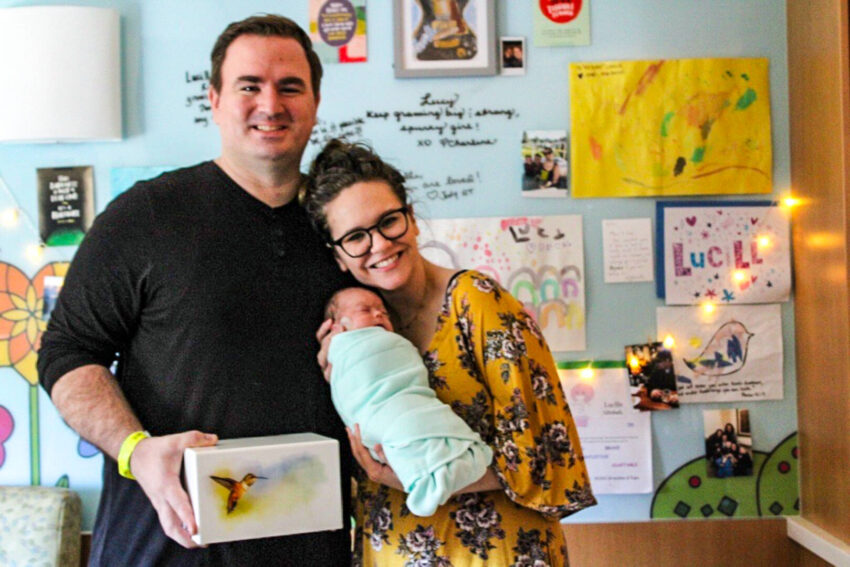 A woman holds an infant and stands next to a man. They both smile in front of a bulletin board filled with drawings.