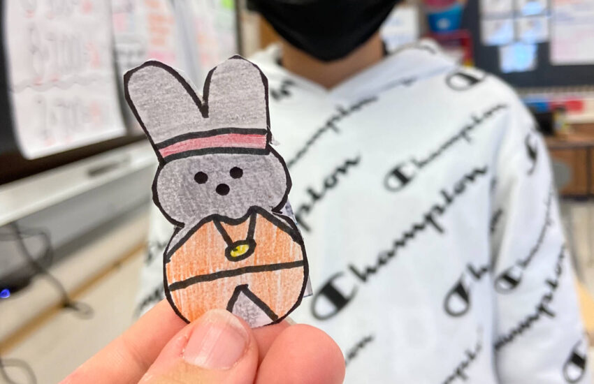 A woman’s hand holds a cutout and hand-colored drawing of a gray Peep bunny wearing an orange cape.