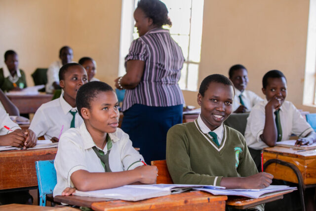 High school students in uniforms listen to their teacher in a classroom. Among them, one girl smiles at the camera.