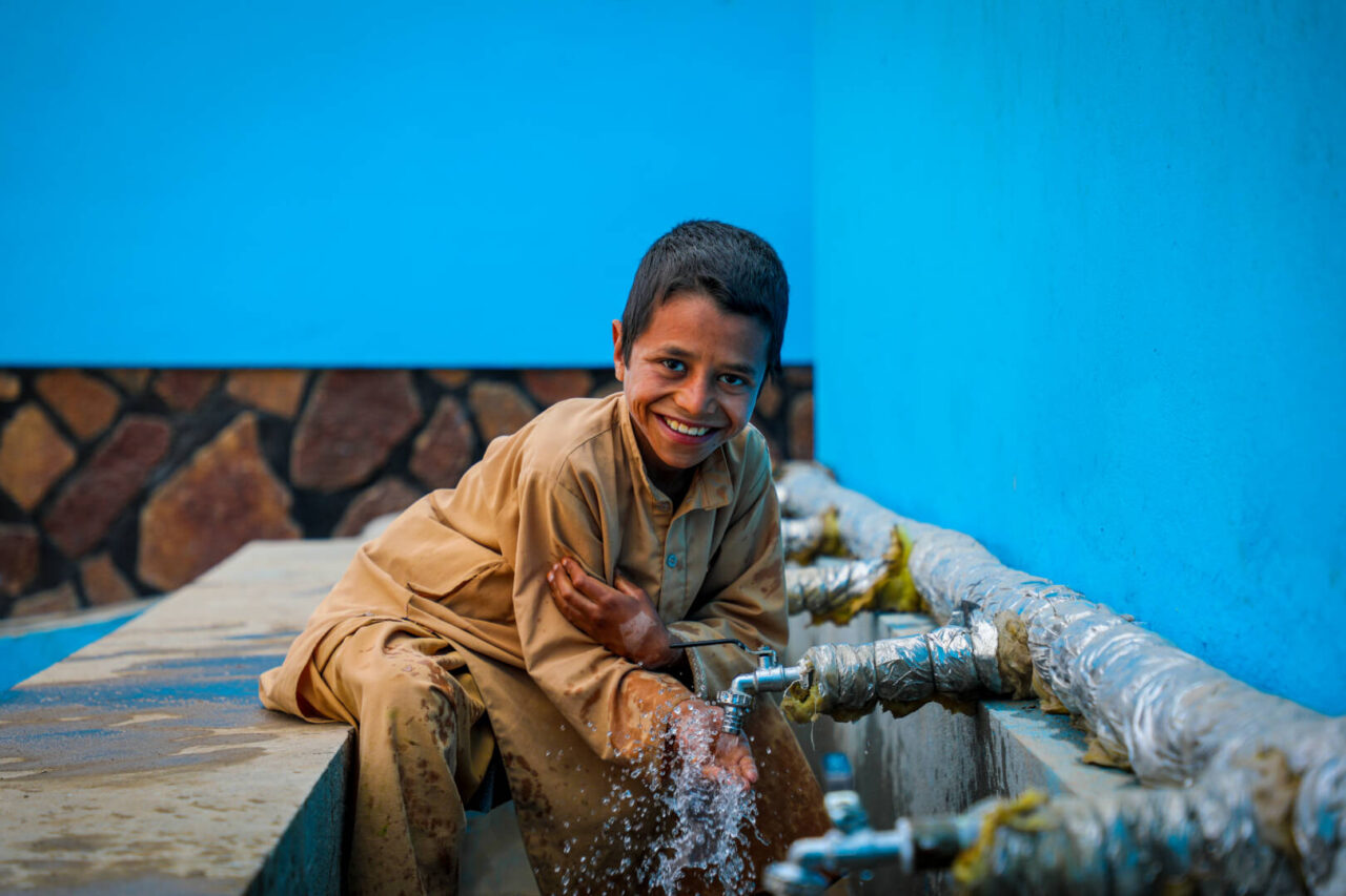 An Afghan boy sits on a cement ledge, water running from a faucet onto his hands. He gazes at the camera with a joyful smile.