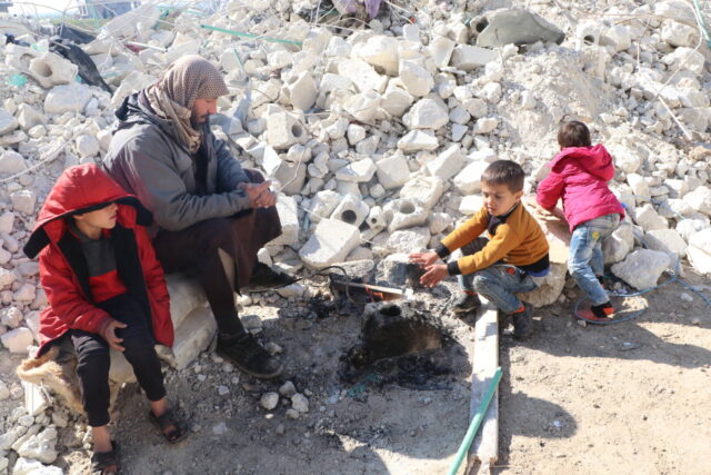 A man and three children sit on the rubble of a collapsed building around a small fire.