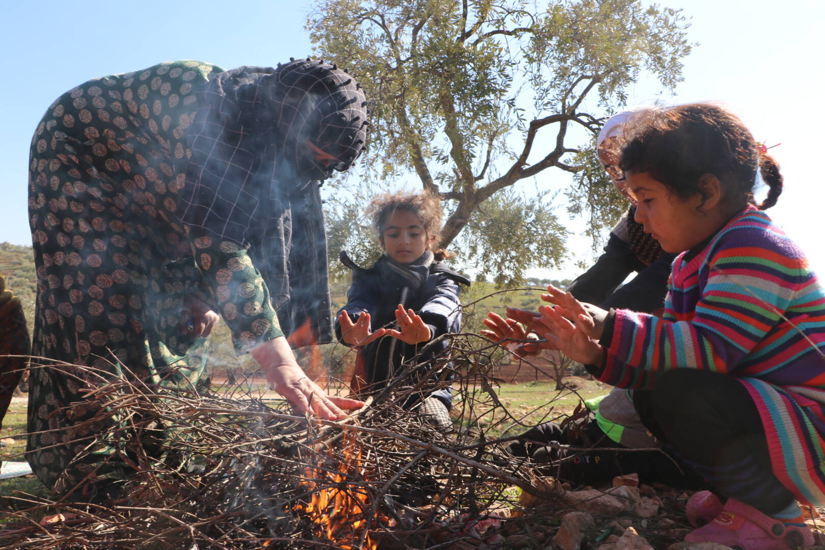 A mother bends down to press her hand on a pile of sticks to kindle a small fire. Three children sit by warming their hands.