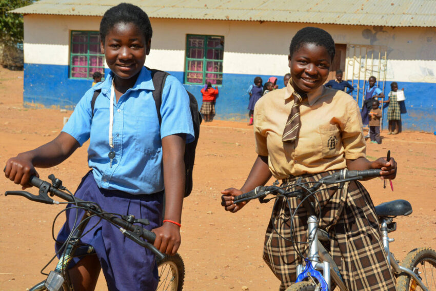 Two Zambian teenage girls in school uniforms stand with their donated bicycles, which help them get to school and do chores safely.