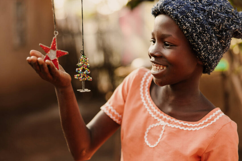 A girl in a winter cap and a pink shirt smiles and lifts her hand to cradle beaded Christmas ornaments hanging from strings.