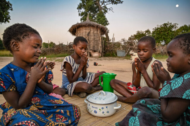 A group of Zambian children pray while sitting around a white pot.