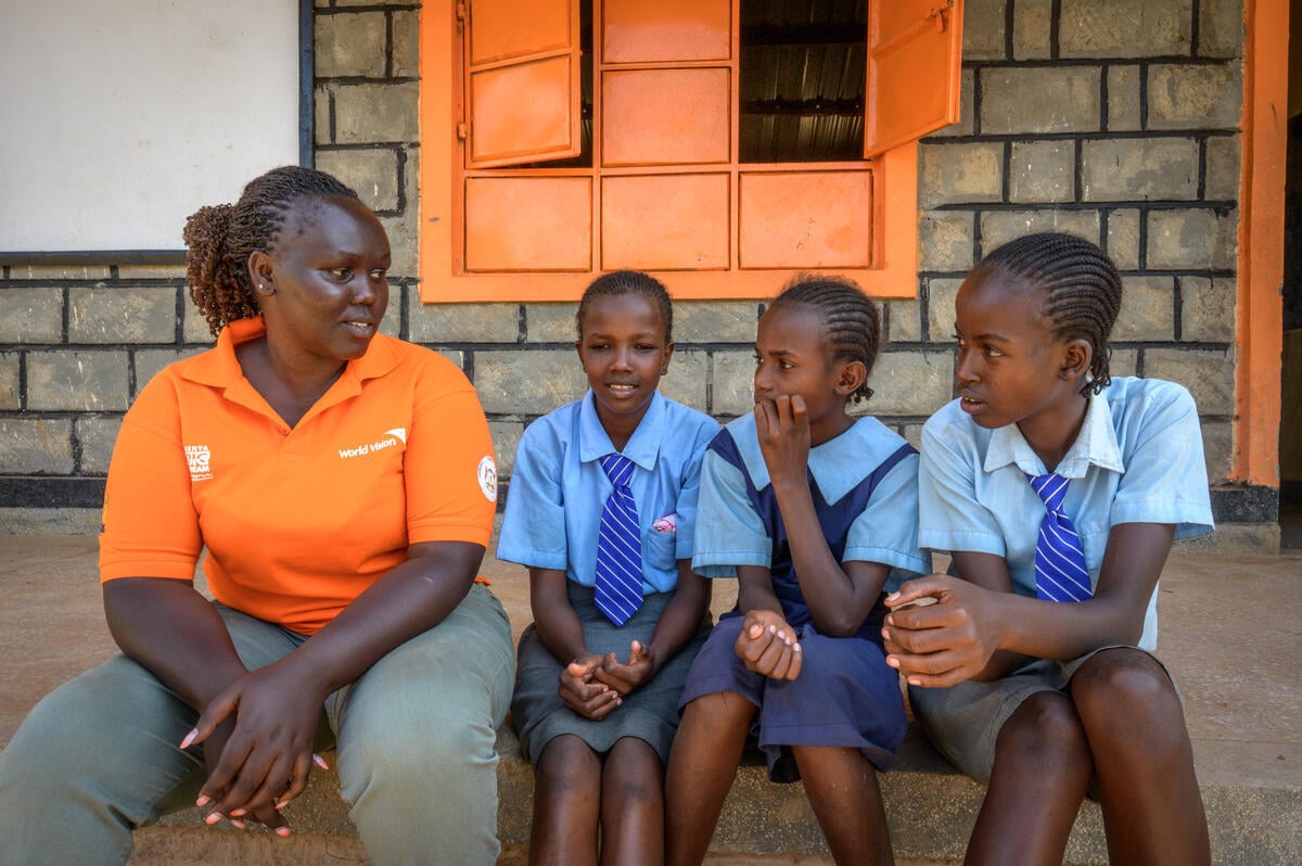 A woman in an orange polo shirt with the World Vision logo sits on the steps with three girls wearing school uniforms.