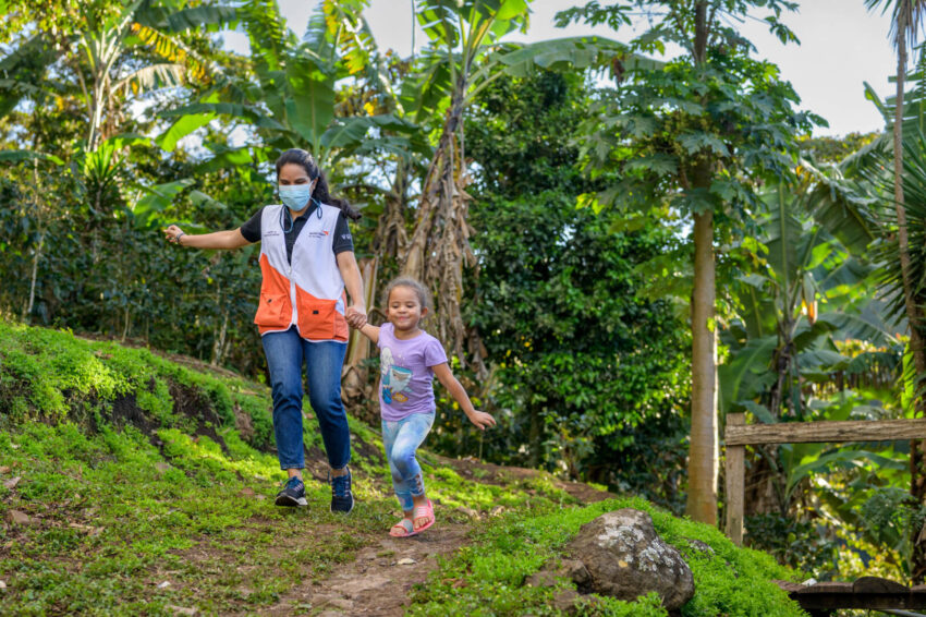 A little girl in a purple T-shirt and a woman in an orange vest run down a green path hand in hand; a palm forest is behind them.