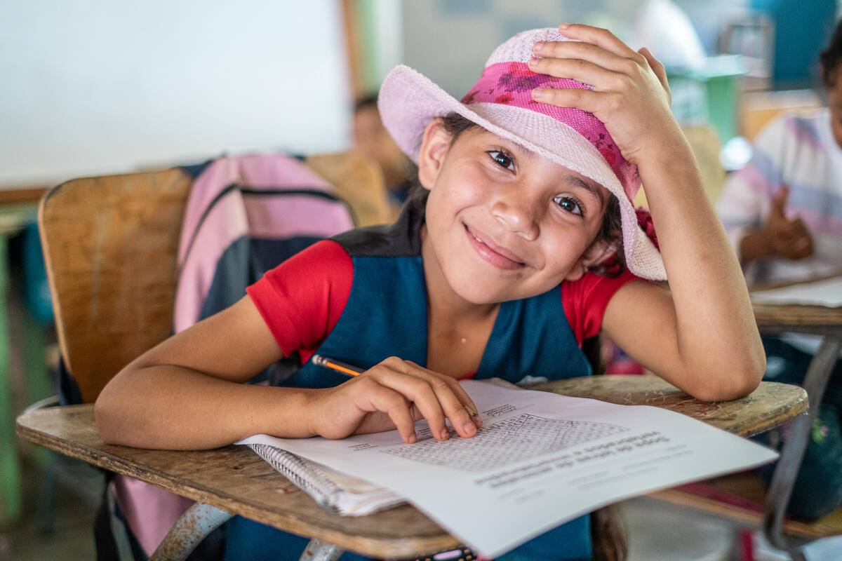 A young girls smiles at a classroom desk with her hand rested on her pink cowboy hat.
