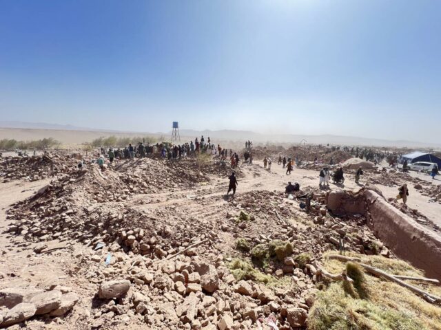 Dust rises from a landscape of rubble and rocks with men standing in the distance amid the aftermath of deadly quakes.