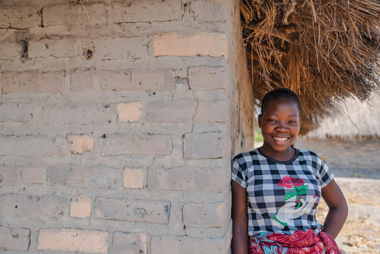 A smile fills a young girl's face as she stands next to a house made of mud and bricks.