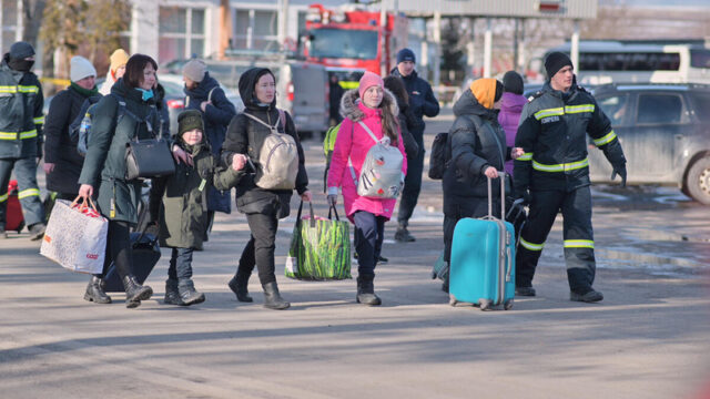 Men in reflective gear guide a group of women and children bundled in winter clothing walk with suitcases and backpacks.
