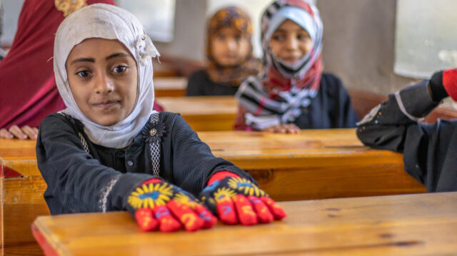 A girl in a white head covering with a faint smile looks at the camera. She wears bright-colored gloves in a classroom setting.