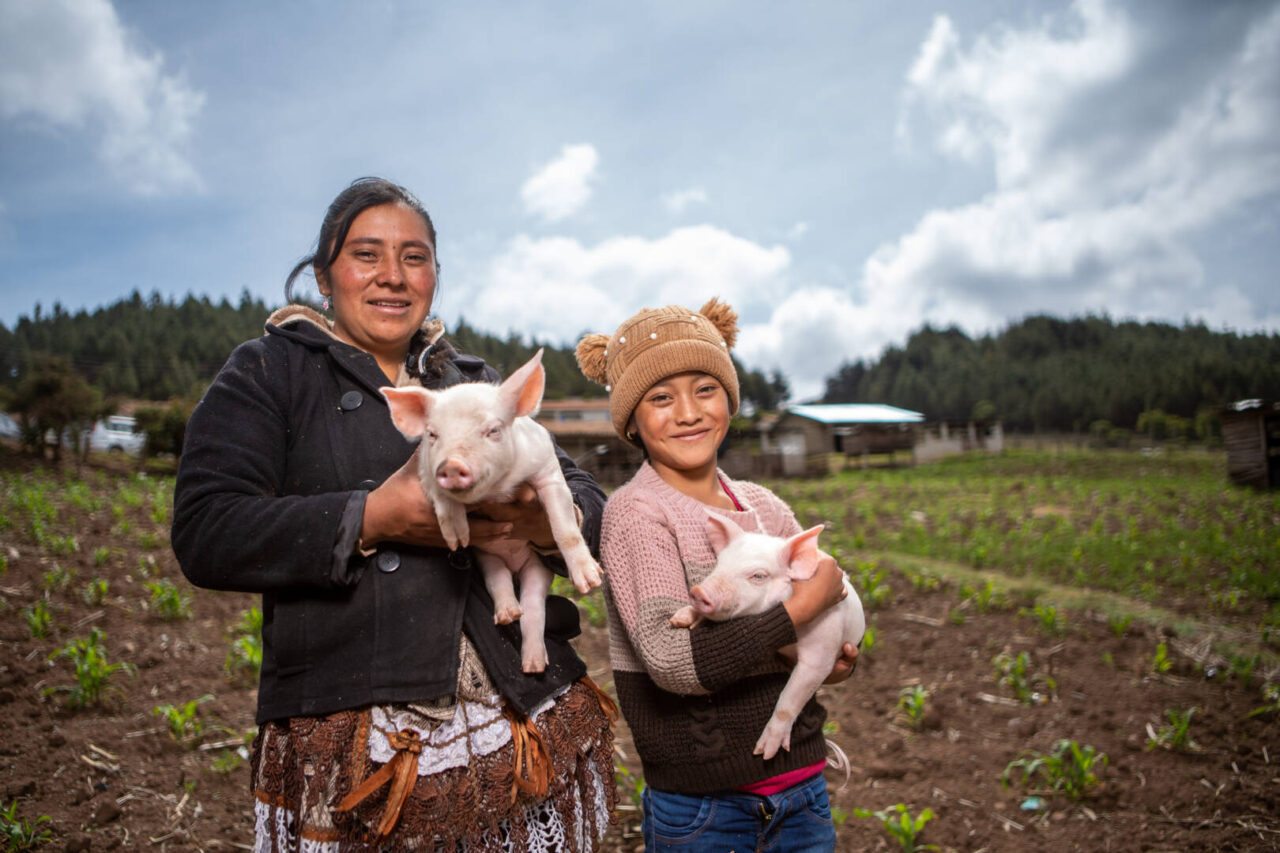 A mother and daughter smile as they pose with their adorable piglets in front of their home.