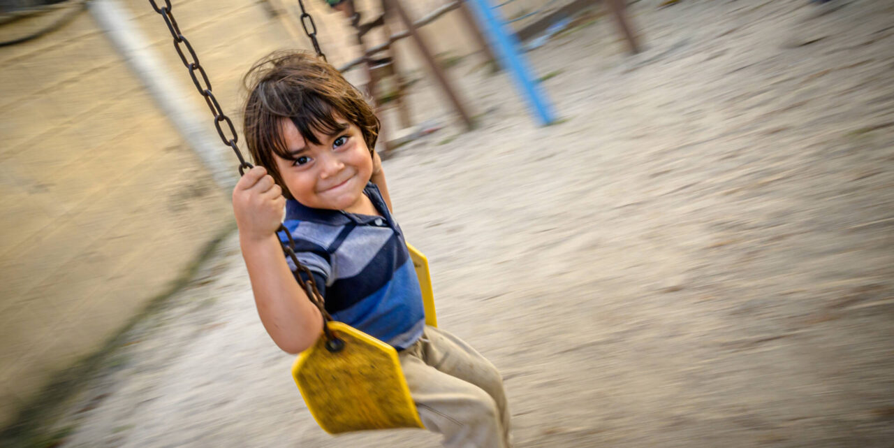 A boy with brown hair sits on a swing in Honduras. He is looking at the camera with a big grin.