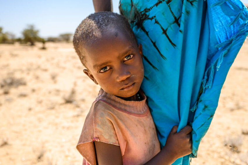 A young Kenyan girl whose family lives in poverty holds her mother’s bright blue skirt. She stands on parched land and looks into the camera.