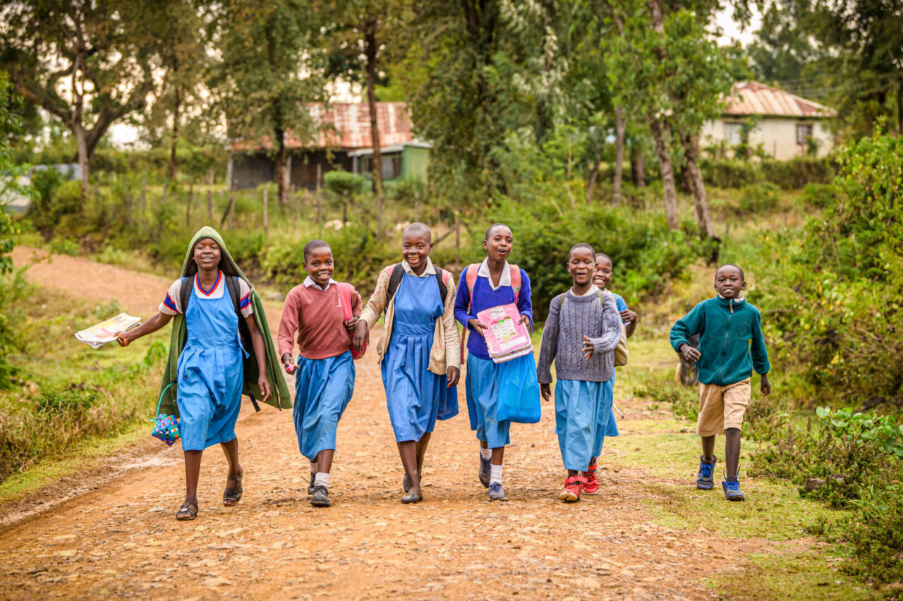 Seven children dressed in school uniforms rush along a dirt road on their way home from school in Kenya.