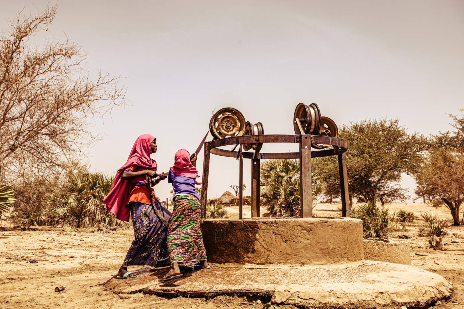 Two girls wearing pink head scarves pull ropes to draw water from an open well.