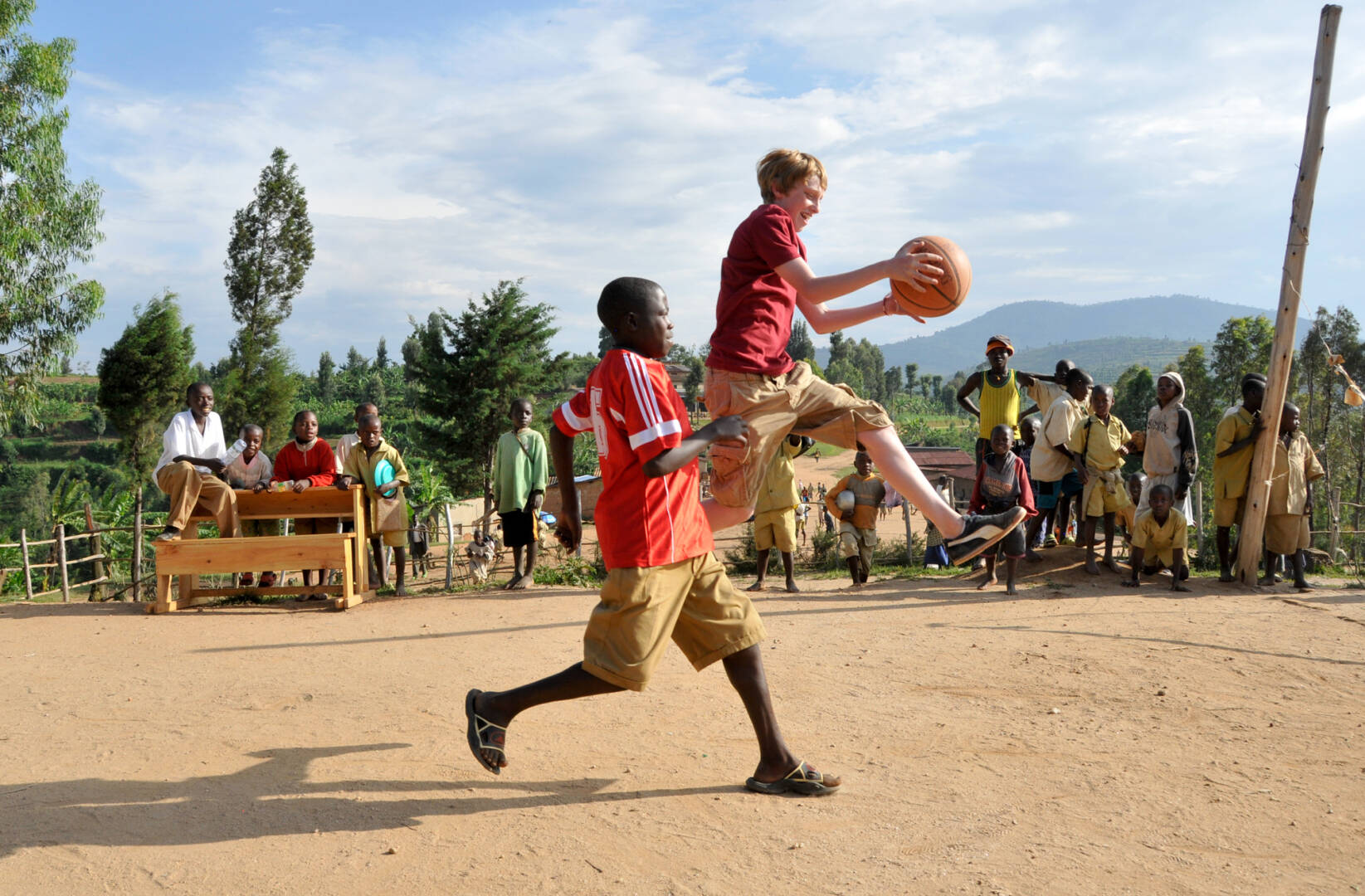 Two boys play basketball on a dirt court while being watched by a group of spectators.