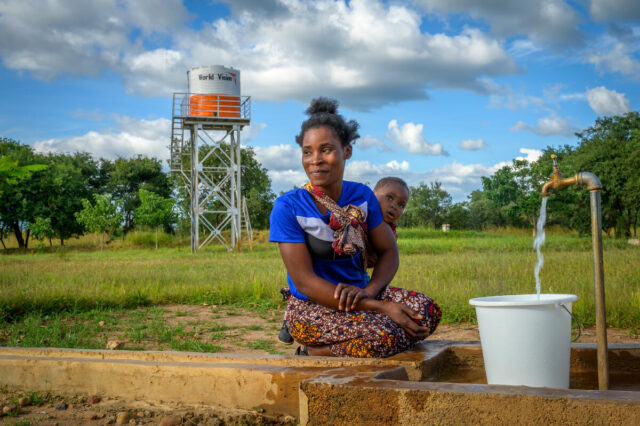 A woman smiles with a bucket of water, a baby tied to her back. A water tower with the World Vision logo is seen in the distance.