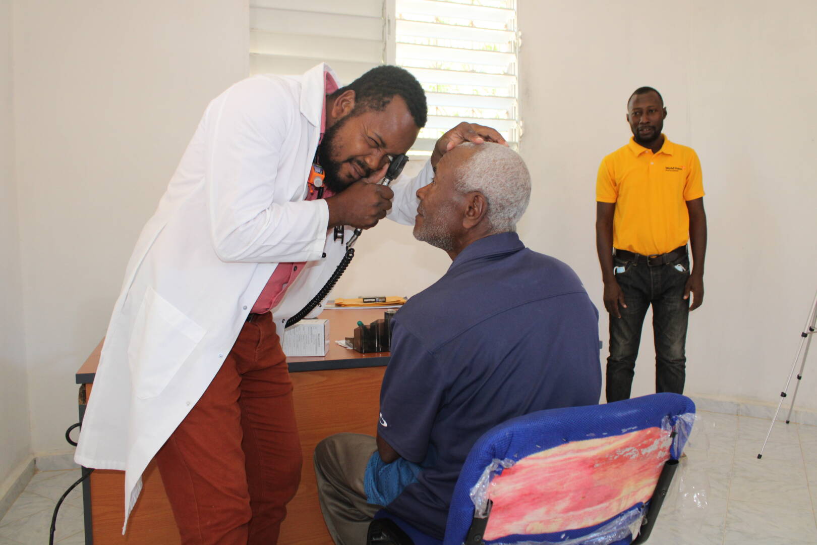 A man in a white lab coat uses a small black device to look into the eyes of an older seated man.