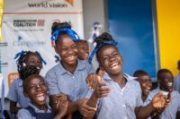Haitian girls in school uniforms and wearing blue hair ribbons laugh and clap joyfully.