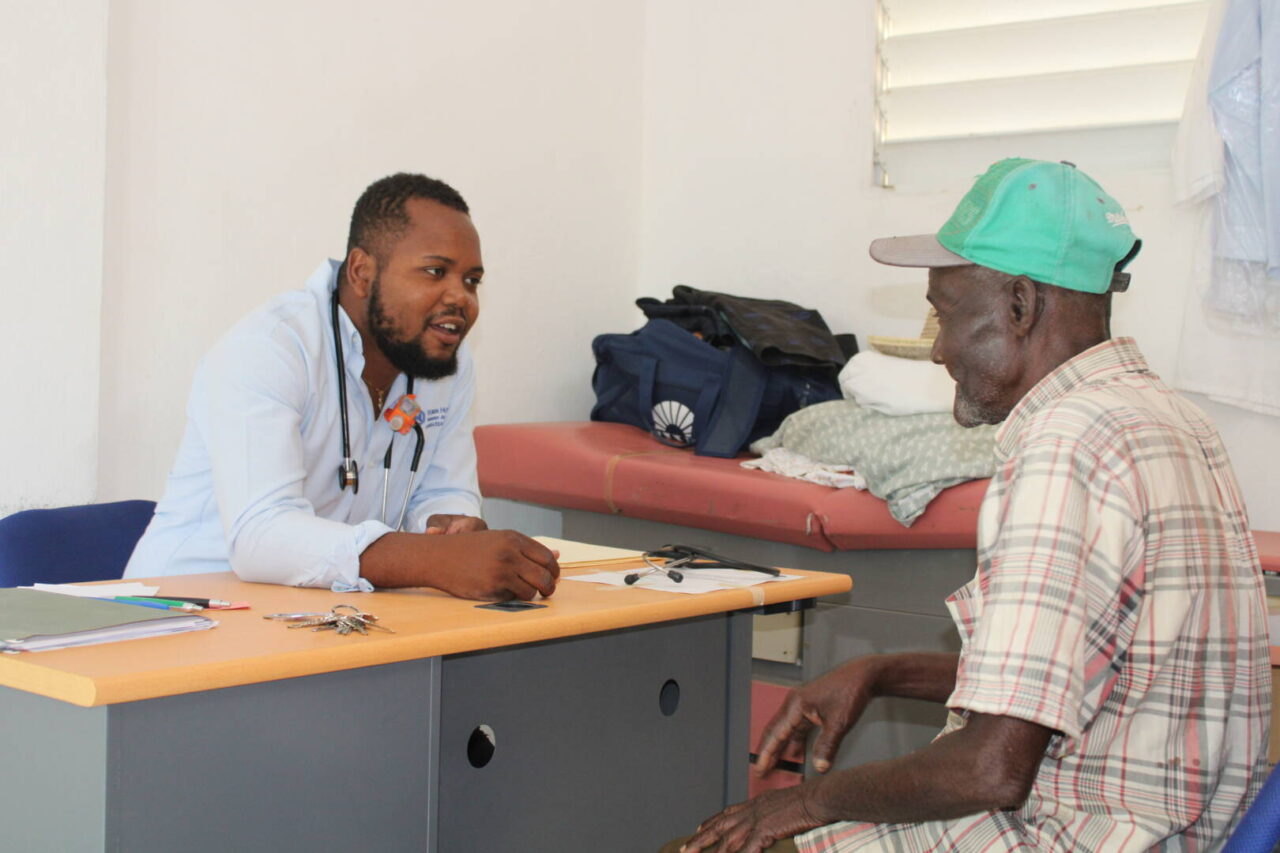 A man wearing a white shirt and stethoscope around his neck sits behind a desk and talks to a man in a Haitian hospital.
