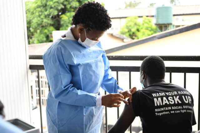 Healthcare worker wearing PPE in Zambia gives a vaccine to a man wearing a black T-shirt that reads “WASH YOUR HANDS. MASK UP.”