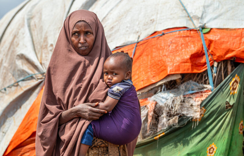 A woman wearing a veil stands holding a child on her hip. Behind them is a shelter made of branches and tarp.