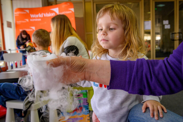 A little girl looks at dry ice in a cup her grandmother holds. Behind her is a World Vision banner in the play area.