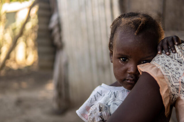 A young girl looks over her mother’s shoulder and into the camera.