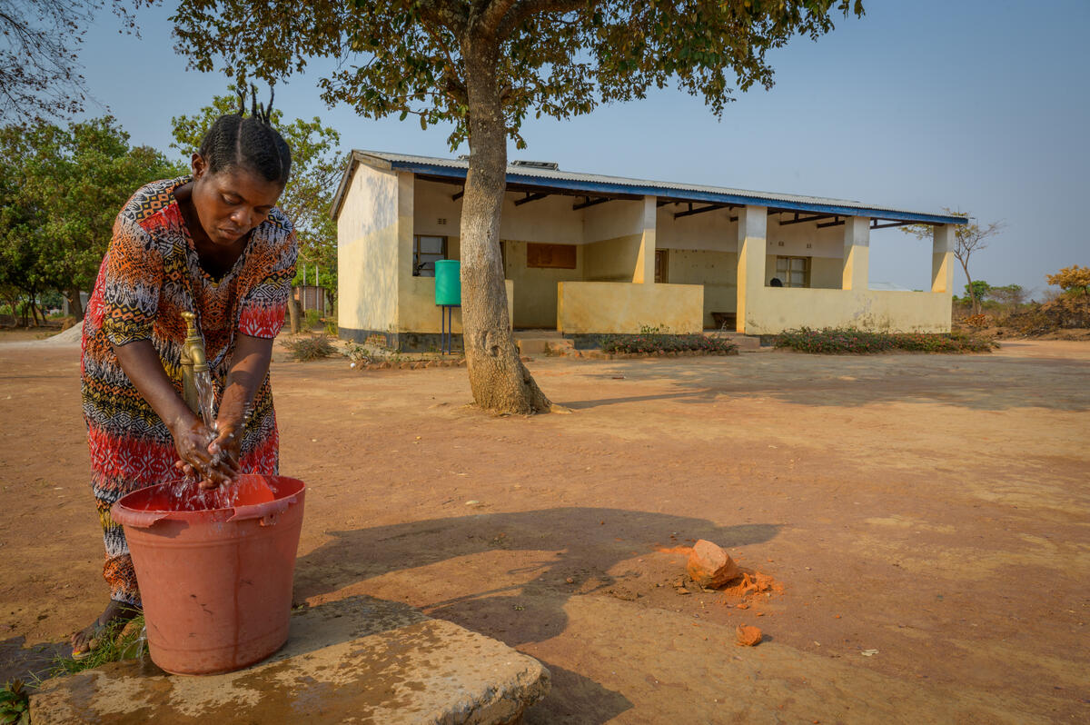 In front of a concrete building, a Zambian woman rinses her hands in water pouring from a tap into a bucket.