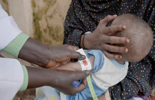 A young child sits on a woman’s lap while the circumference of the child’s arm is measured for malnutrition. The tape shows red.