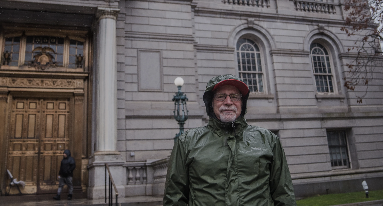 In front of a neoclassical building, a man in a green raincoat stands with his hands in the front pockets of his jeans.