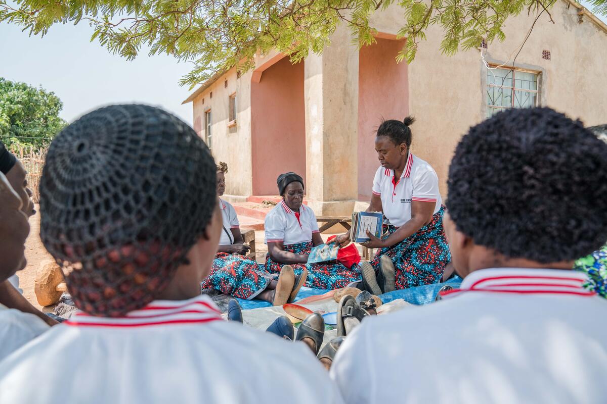 A group of women sit in a circle, one distributing notebooks. All wear matching colorful skirts and white collared shirts.