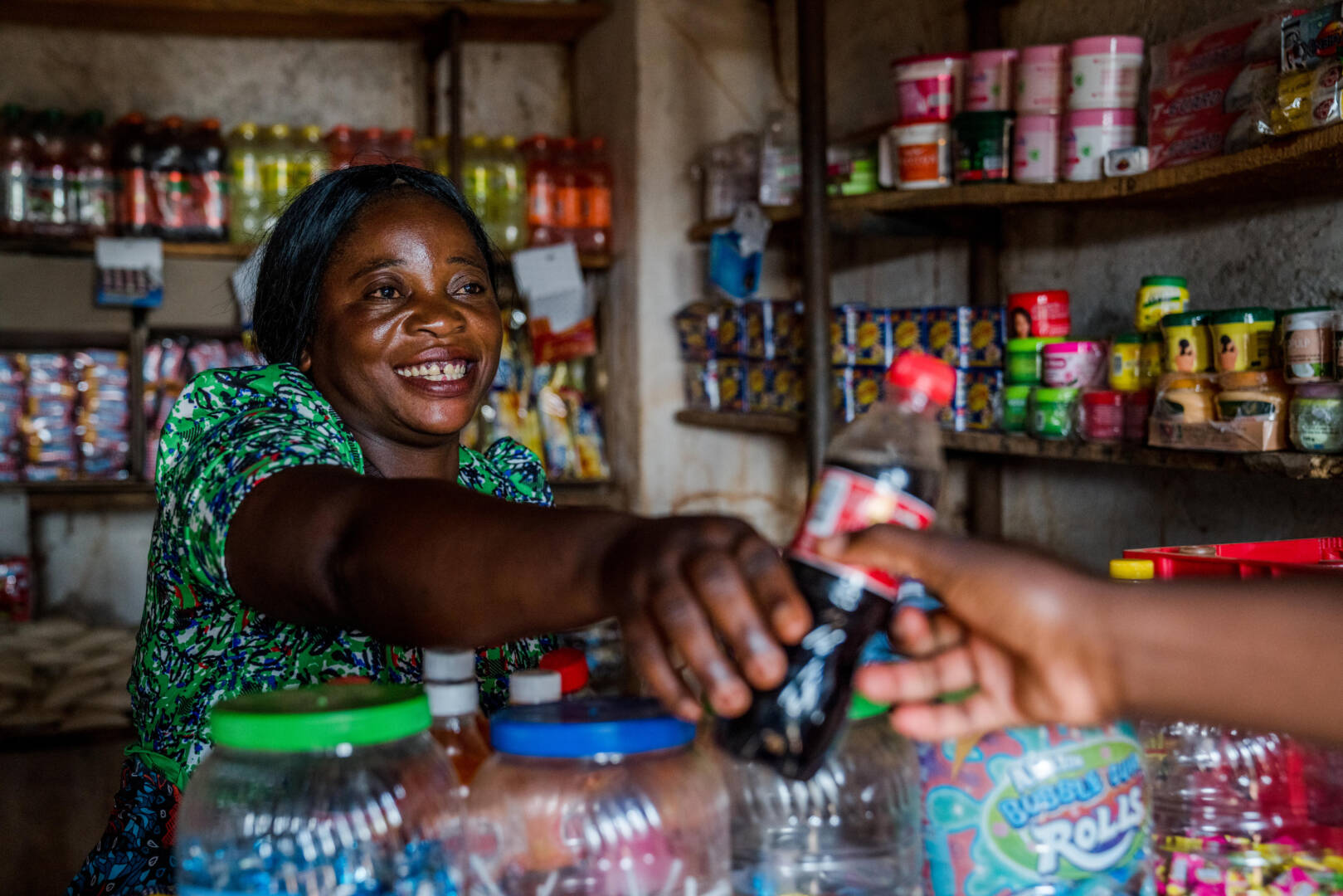 Inside a small shop, a smiling woman hands a drink to a customer over a counter.